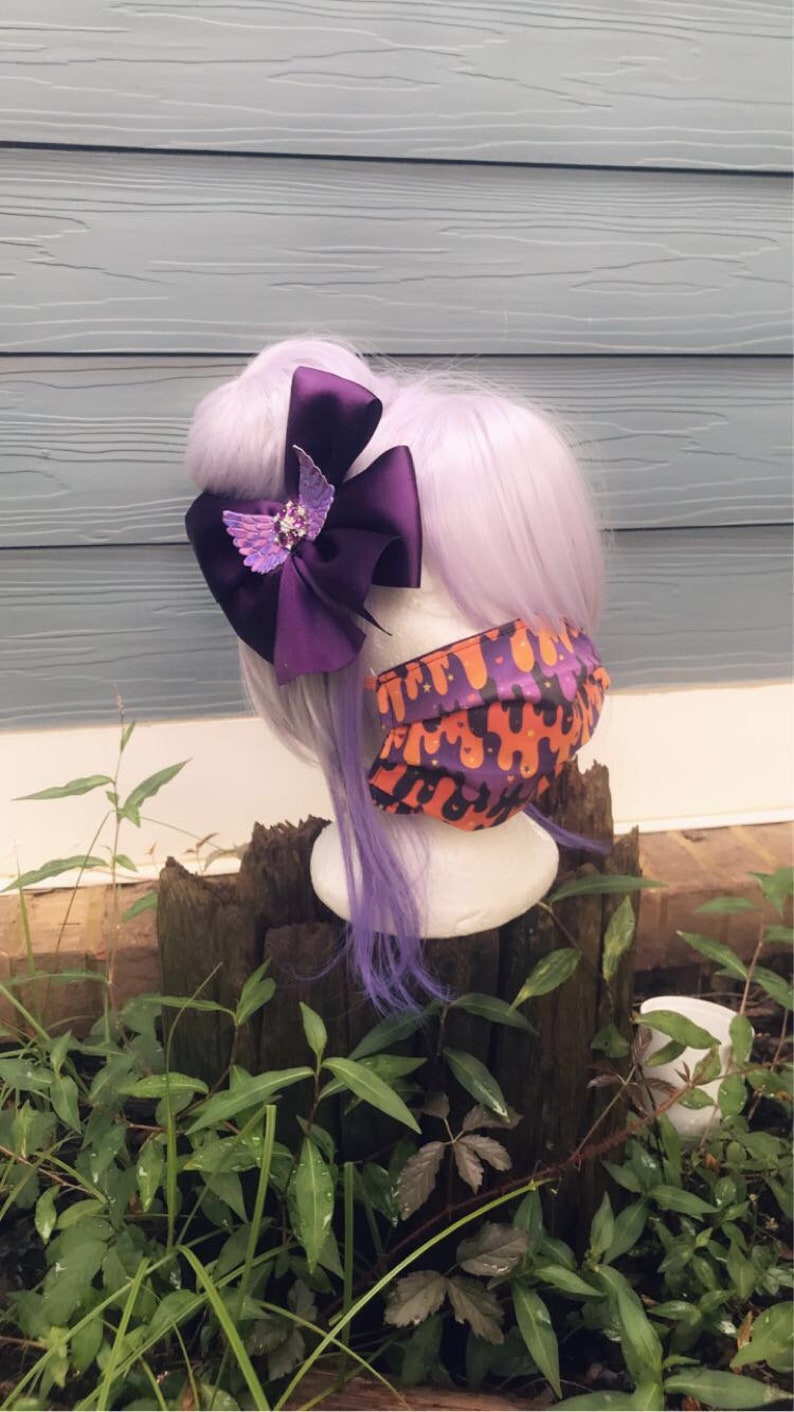 May include: A purple and orange patterned face mask with a purple bow with a sparkly butterfly accent in the center. The bow is attached to a purple wig.