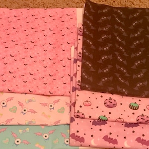 May include: A stack of eight fabric squares with various Halloween-themed patterns. The patterns include bats, stars, pumpkins, and eyeballs. The fabrics are in shades of pink, brown, and blue.