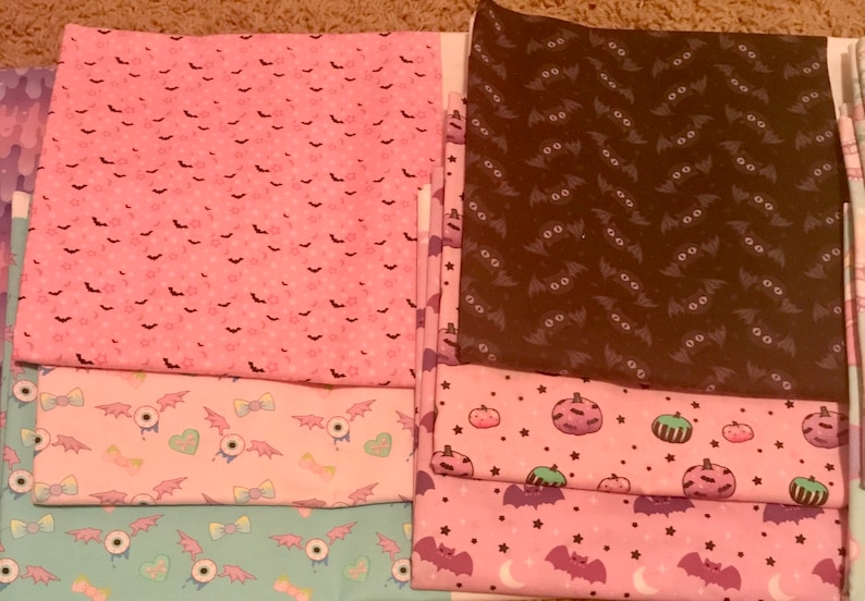 May include: A stack of eight fabric squares with various Halloween-themed patterns. The patterns include bats, stars, pumpkins, and eyeballs. The fabrics are in shades of pink, brown, and blue.