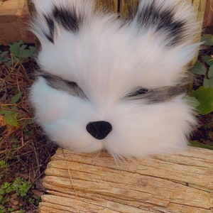 May include: A plush white raccoon head with black eyes and a black nose. The raccoon head is resting on a weathered wooden log.