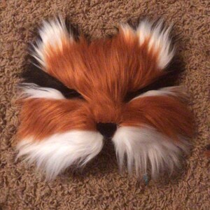 May include: A red and white furry fox mask with black eyes. The mask is made of soft, plush fabric and has a realistic fox face.
