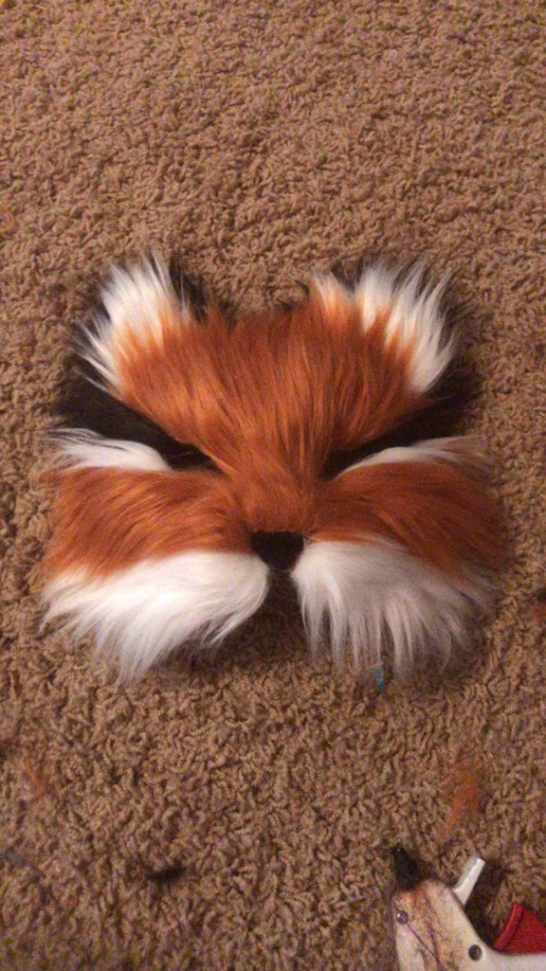May include: A red and white furry fox mask with black eyes. The mask is made of soft, plush fabric and has a realistic fox face.