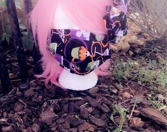 May include: A pink wig with a black and purple Halloween-themed face mask and a matching headband with a bow. The mask has a variety of Halloween-themed designs, including ghosts, pumpkins, and bats.