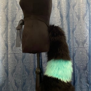 Puede incluir: Una cola de piel sintética negra con una banda rayada turquesa y negra. La cola está unida a un maniquí negro sobre un soporte de madera.