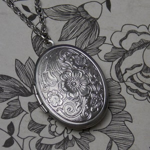 May include: A silver locket necklace with a floral design. The locket is oval shaped and has a hinged opening.