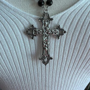 May include: A silver cross pendant necklace with intricate floral designs. The cross is large and ornate, with a black beaded chain.