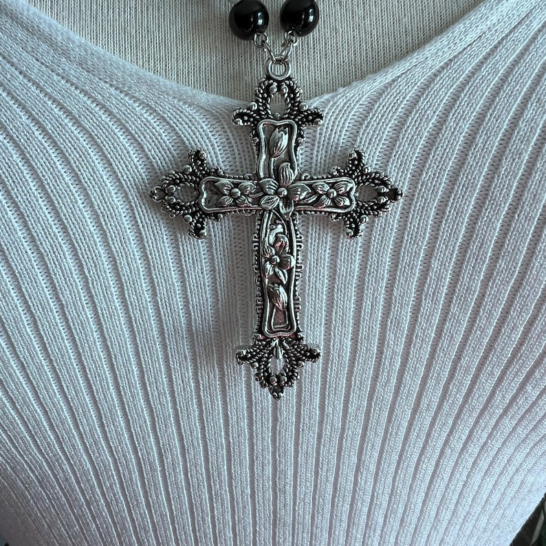 May include: A silver cross pendant necklace with intricate floral designs. The cross is large and ornate, with a black beaded chain.