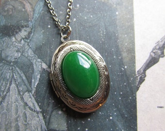 Puede incluir: Un collar con un medallón de plata adornado con una piedra ovalada verde. El medallón tiene un diseño detallado alrededor del borde.