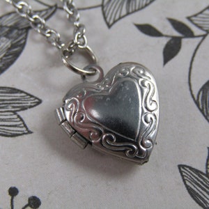 May include: A silver heart-shaped locket necklace with a delicate chain and an intricate design. The locket has a hinged opening and a detailed floral pattern.