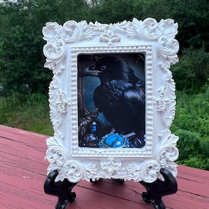 May include: A white ornate frame with a black crow perched on a pile of blue and black gemstones. The frame is on a black stand and the background is a wooden surface.