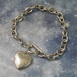 May include: A silver chain bracelet with a heart-shaped locket pendant. The chain is a curb style and the locket has a detailed design.