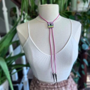 May include: A pink bolo tie with a square pendant featuring a black cat design. The tie has a braided cord and silver-coloured tips. The cat is set against a colourful background. The bolo tie is displayed on a white mannequin.