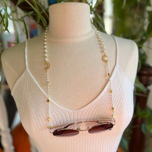 May include: A white pearl and gold chain with crescent moon and star charms. The chain is being used as a glasses chain and is holding a pair of brown sunglasses.