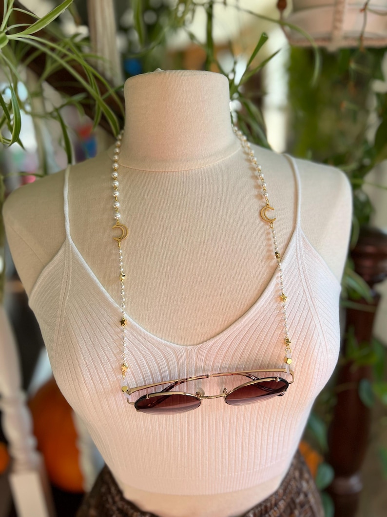 May include: A white pearl and gold chain with crescent moon and star charms. The chain is being used as a glasses chain and is holding a pair of brown sunglasses.