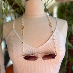 May include: A white pearl and gold chain with crescent moon and star charms. The chain is being used as a glasses chain and is holding a pair of brown sunglasses.