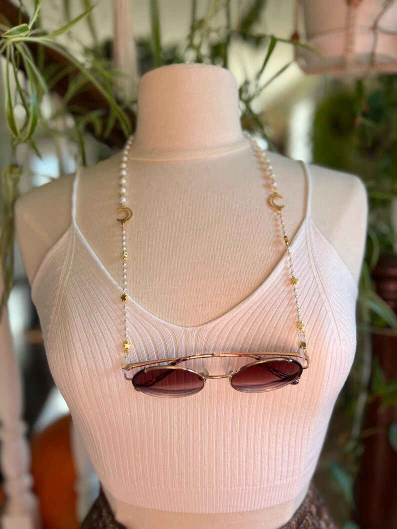 May include: A white pearl and gold chain with crescent moon and star charms. The chain is being used as a glasses chain and is holding a pair of brown sunglasses.