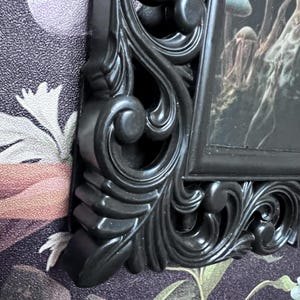 May include: Black ornate picture frame with a detailed, intricate design. The frame is made of plastic and has a dark, almost black finish.