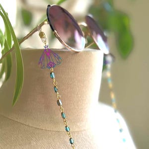 May include: A pair of round sunglasses with a gold-toned chain featuring small iridescent blue beads and a moth-shaped charm. The moth charm displays a gradient of purple and blue hues with thin, dark outlines, attached to the glasses by clear loops.
