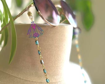 May include: A pair of round sunglasses with a gold-toned chain featuring small iridescent blue beads and a moth-shaped charm. The moth charm displays a gradient of purple and blue hues with thin, dark outlines, attached to the glasses by clear loops.