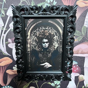 May include: A black ornate frame with a gothic-style print of a woman with a sugar skull makeup design. The woman is wearing a black dress with gold accents and a crown of roses.