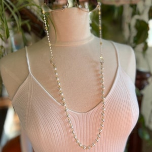 May include: A white pearl and gold chain with a clasp on each end. The chain is worn as a glasses chain, with the ends looped around the arms of a pair of sunglasses.