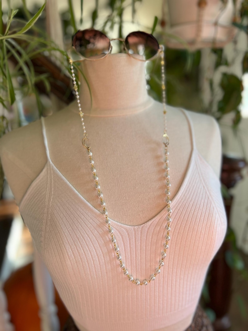 May include: A white pearl and gold chain with a clasp on each end. The chain is worn as a glasses chain, with the ends looped around the arms of a pair of sunglasses.