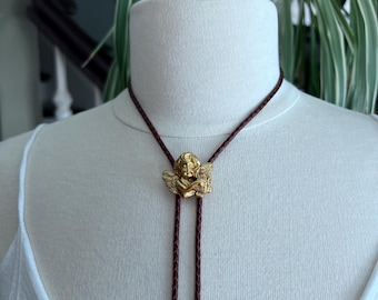 May include: A brown leather bolo tie with a gold cherub pendant. The cherub is holding a heart in its hands.