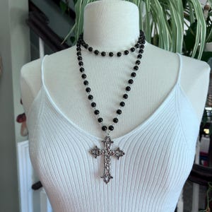 May include: A black beaded necklace with a silver cross pendant. The necklace is layered and has a simple design.