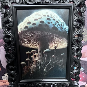May include: A framed print of a large, blue and white mushroom with a dark brown stem and a cluster of smaller mushrooms at the base. The mushrooms are set against a dark background.