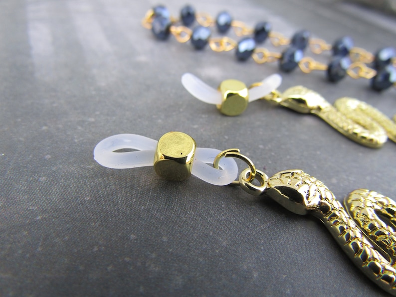May include: Two gold-toned eyeglass chain ends. One end features a clear plastic loop with a gold-toned cube. The other end features a gold-toned snake charm.