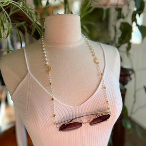 May include: A white pearl and gold chain with crescent moon and star charms. The chain is being used as a glasses chain and is holding a pair of brown sunglasses.