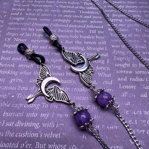 May include: A pair of silver-toned eyeglass chains featuring a crescent moon and bird charm design. Each chain includes a round purple gemstone bead, a small flower-shaped metal accent, and black rubber loops for attaching to the frames of eyeglasses.