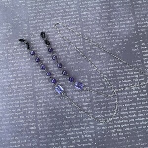 May include: Two silver chains with amethyst beads and a silver charm with a raven design. The chains are draped over a book with text about a raven.