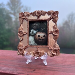 May include: A small, ornate gold-colored frame with a picture of a young girl holding a carved pumpkin. The frame is on a clear plastic stand.