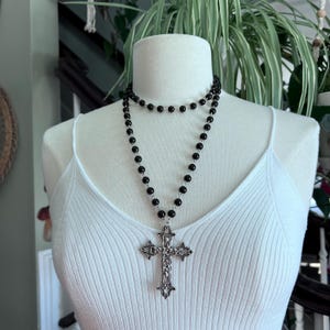 May include: A black beaded necklace with a large silver cross pendant. The necklace is layered and has a simple design.