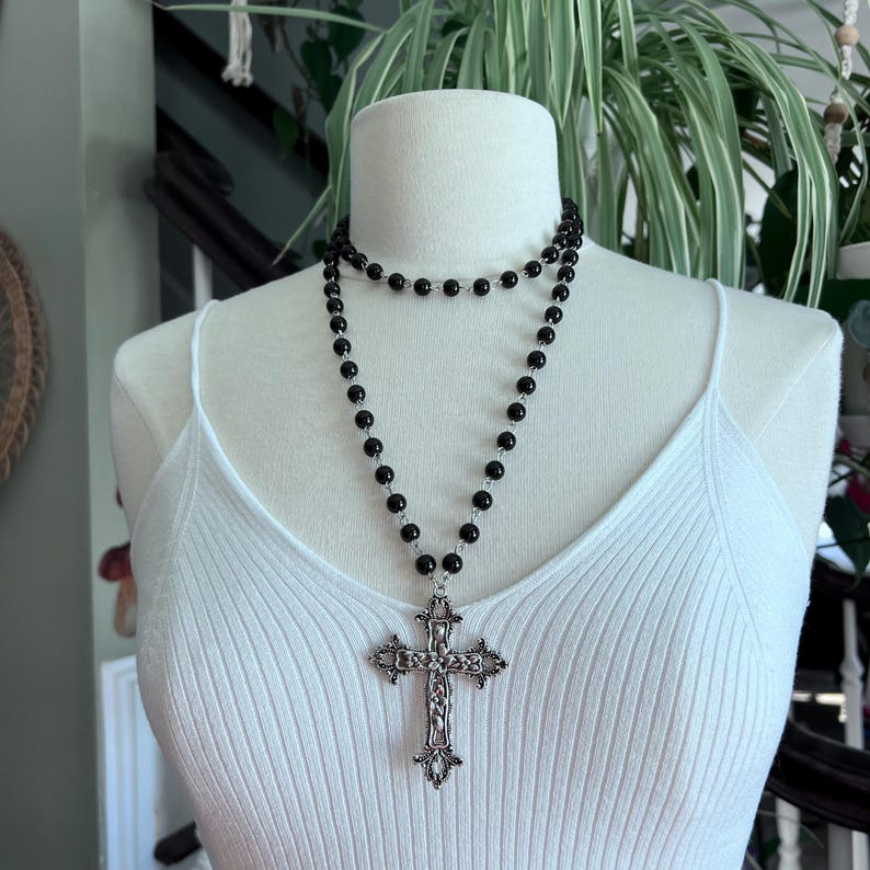 May include: A black beaded necklace with a large silver cross pendant. The necklace is layered and has a simple design.