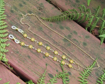 May include: A gold chain with yellow flower accents, designed to hold glasses. The chain is laying on a wooden surface.