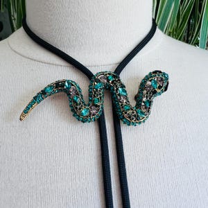 May include: A bolo tie featuring a snake-shaped pendant encrusted with numerous small, sparkling emerald green and dark grey crystals set in a gold-toned metal frame, suspended from a simple, smooth black cord that hangs down in two parallel lines.