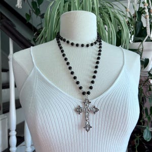 May include: A black beaded necklace with a large silver cross pendant. The necklace is layered and has a choker style.