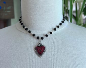 May include: A black beaded necklace with a red heart pendant. The heart pendant has a silver frame with a rose at the top and a textured red center. The necklace is displayed on a white mannequin.