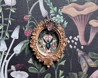 May include: Ornate gold-colored framed pendant with a black beetle illustration. The pendant has a pink gemstone and a silver-colored spider-like design on top. The background is a dark floral pattern with mushrooms and strawberries.