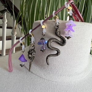 May include: A set of four decorative charms attached to a pink eyeglass frame with translucent silicone rings and gold-tone beads. The charms include a purple flower, a metal bird skull, a blue mushroom, a metal snake, and a stacked purple flower.