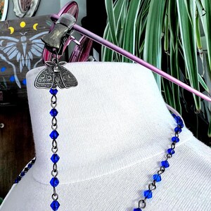 May include: A silver-toned moth-shaped eyeglass holder with a chain of blue beads. The eyeglass holder is attached to a pair of pink spectacles. The necklace is made of blue beads.