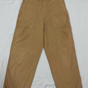 Könnte beinhalten: Beige Hose mit weitem Bein, zwei Vordertaschen und einem Knopfverschluss. Diese hoch taillierte Hose bietet eine entspannte Passform, ideal für den Alltag. Das Material scheint eine robuste Baumwollmischung zu sein.