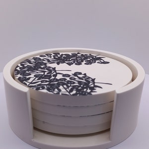 May include: Set of four white ceramic coasters with a black floral design, stacked in a white holder.