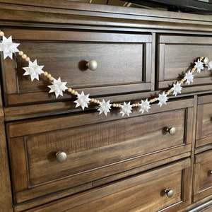 May include: A white paper star garland with wooden beads hangs on a dark brown wooden dresser with multiple drawers. The garland is made of white paper stars and wooden beads.