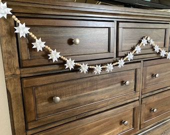 May include: A white paper star garland with wooden beads hangs on a dark brown wooden dresser with multiple drawers. The garland is made of white paper stars and wooden beads.
