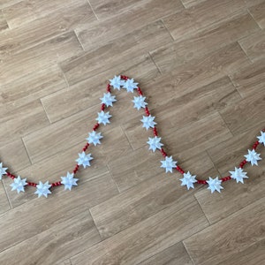 May include: A decorative garland featuring white star-shaped ornaments strung on a red beaded strand. The garland is laid out on a wooden surface, creating a festive and eye-catching display.