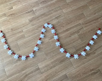May include: A decorative garland featuring white star-shaped ornaments strung on a red beaded strand. The garland is laid out on a wooden surface, creating a festive and eye-catching display.