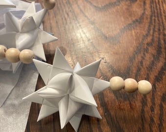 May include: Three white paper stars with wooden beads strung through them. The stars are folded into a three-dimensional shape.